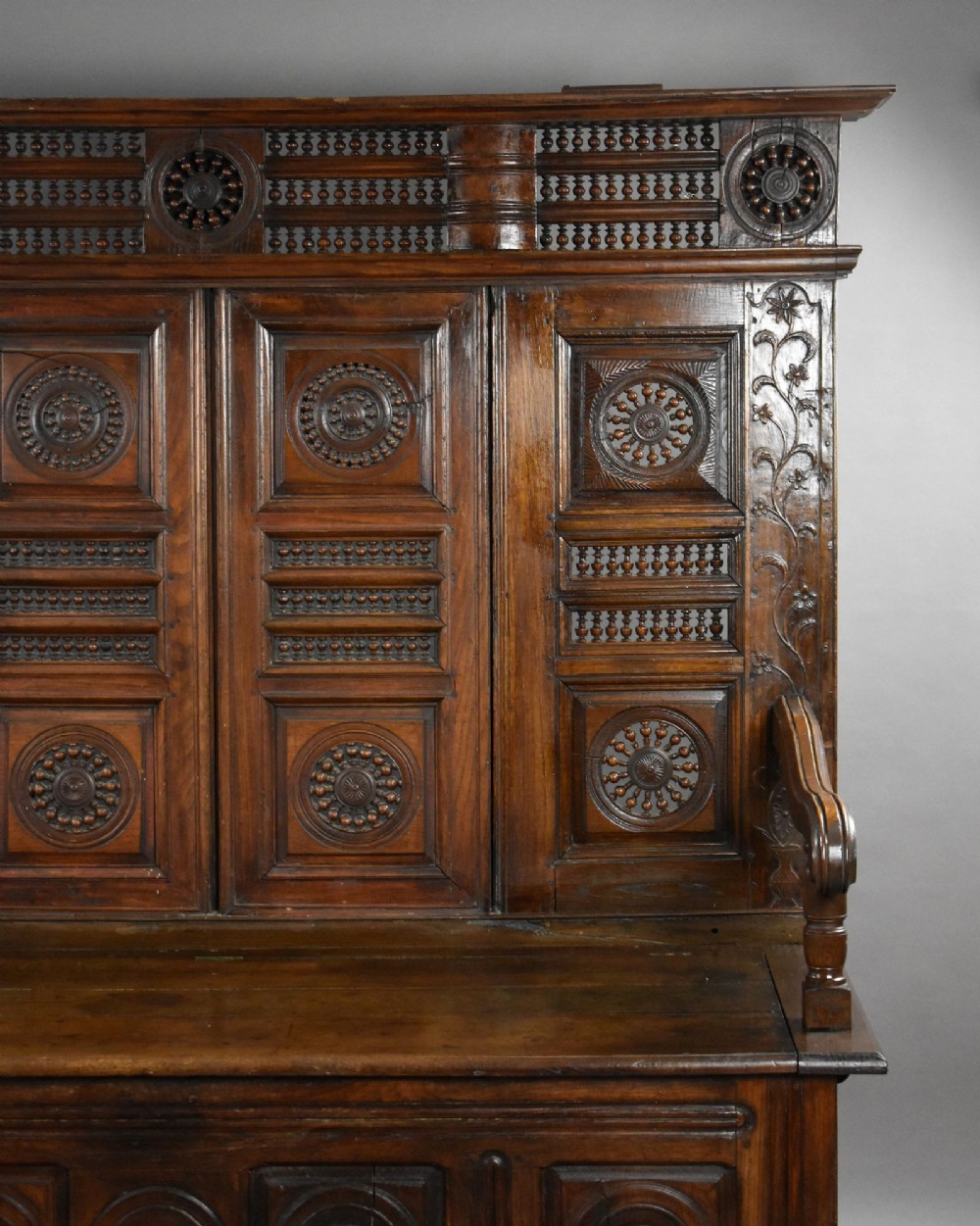 A high-backed dark oak panelled settle featuring geometric roundel carvings, a spindle frieze, scroll arms, and a box seat storage base. View V