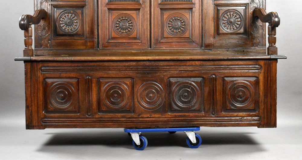 A high-backed dark oak panelled settle featuring geometric roundel carvings, a spindle frieze, scroll arms, and a box seat storage base. View VI