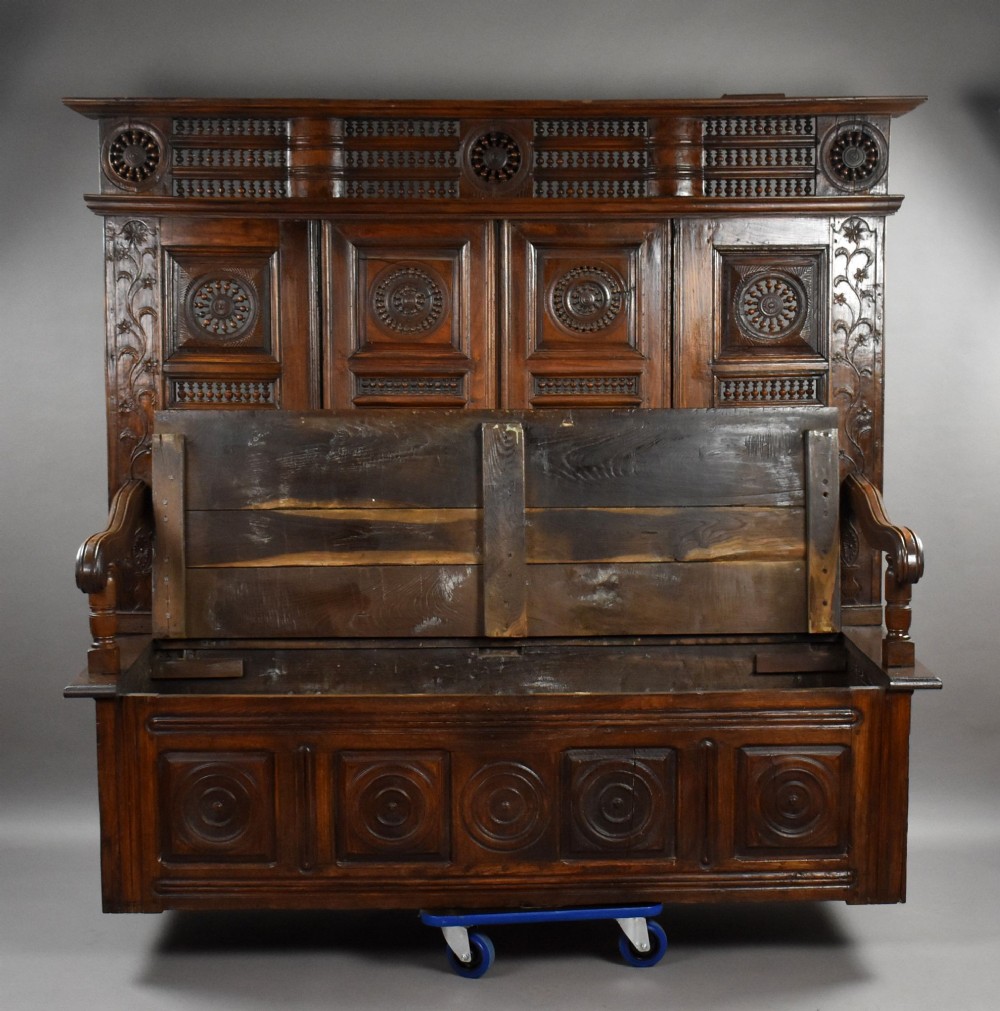 A high-backed dark oak panelled settle featuring geometric roundel carvings, a spindle frieze, scroll arms, and a box seat storage base. View IX