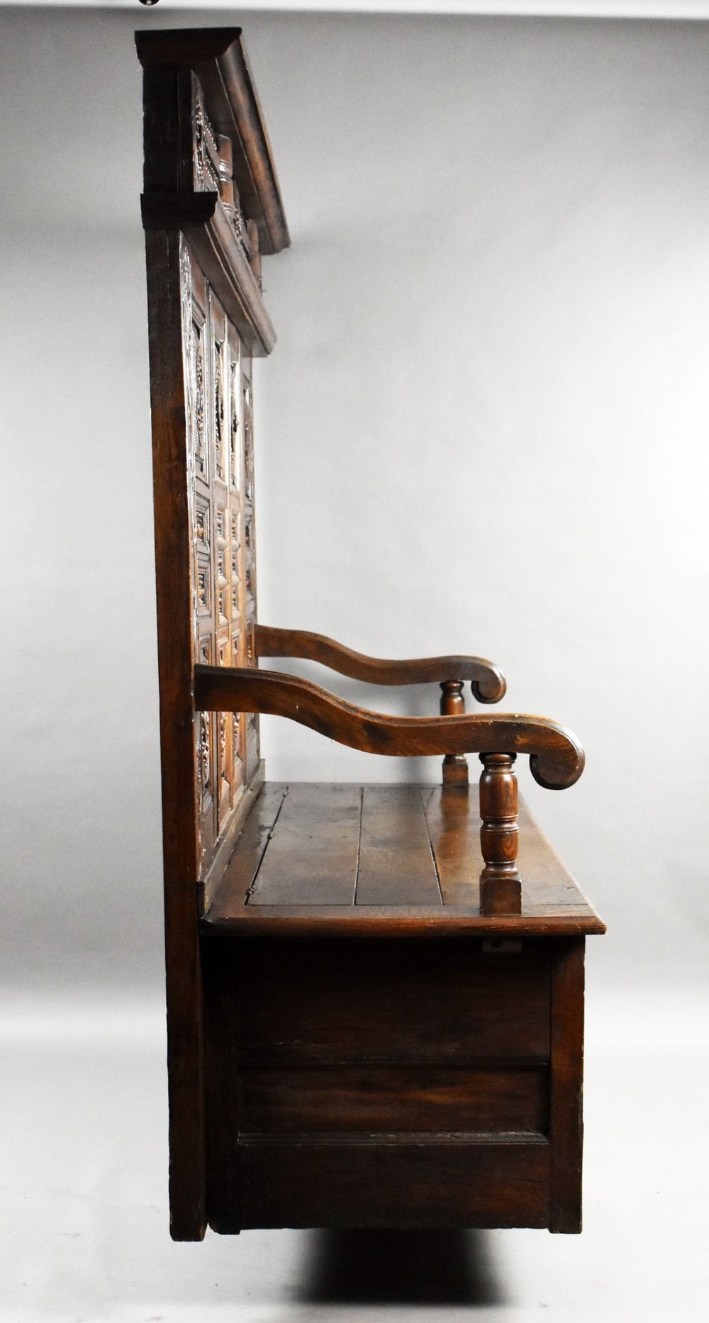 A high-backed dark oak panelled settle featuring geometric roundel carvings, a spindle frieze, scroll arms, and a box seat storage base. View X