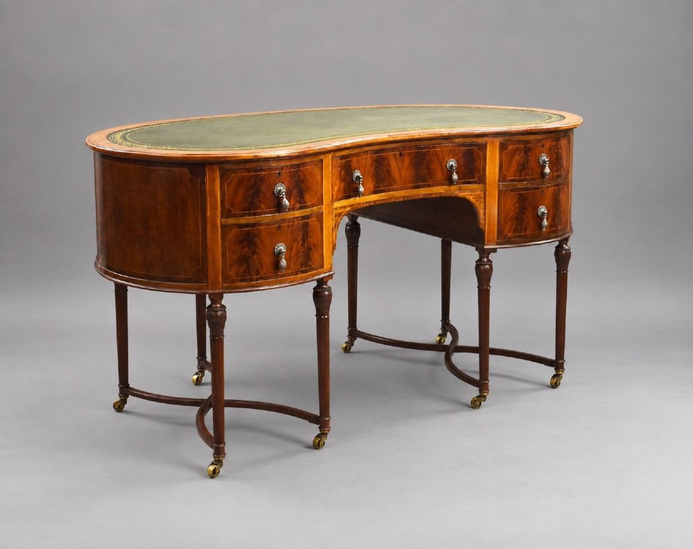 An Edwardian kidney-shaped writing desk crafted from flame mahogany with a green leather writing surface, five drawers, turned fluted legs, and brass castors.