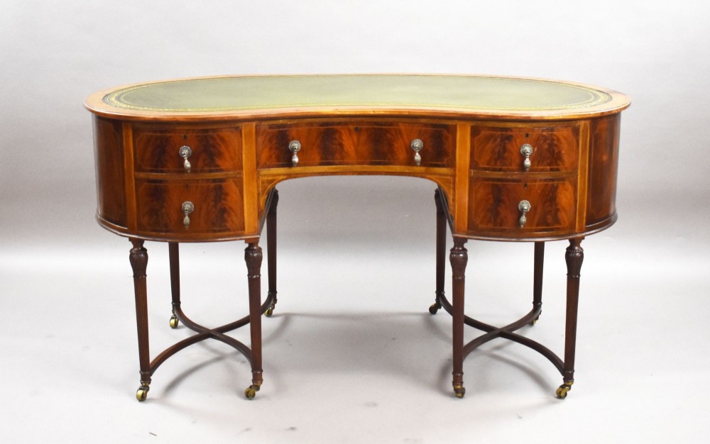 An Edwardian kidney-shaped writing desk crafted from flame mahogany with a green leather writing surface, five drawers, turned fluted legs, and brass castors. View II