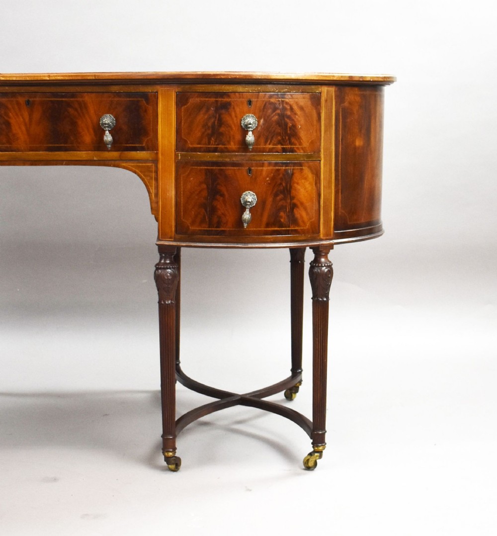 An Edwardian kidney-shaped writing desk crafted from flame mahogany with a green leather writing surface, five drawers, turned fluted legs, and brass castors. View V