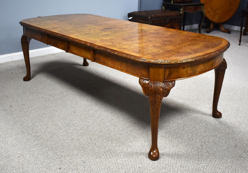 A Queen Anne style burr walnut extending dining table shown with eight matching chairs featuring curved backrests and floral upholstered seats. View II