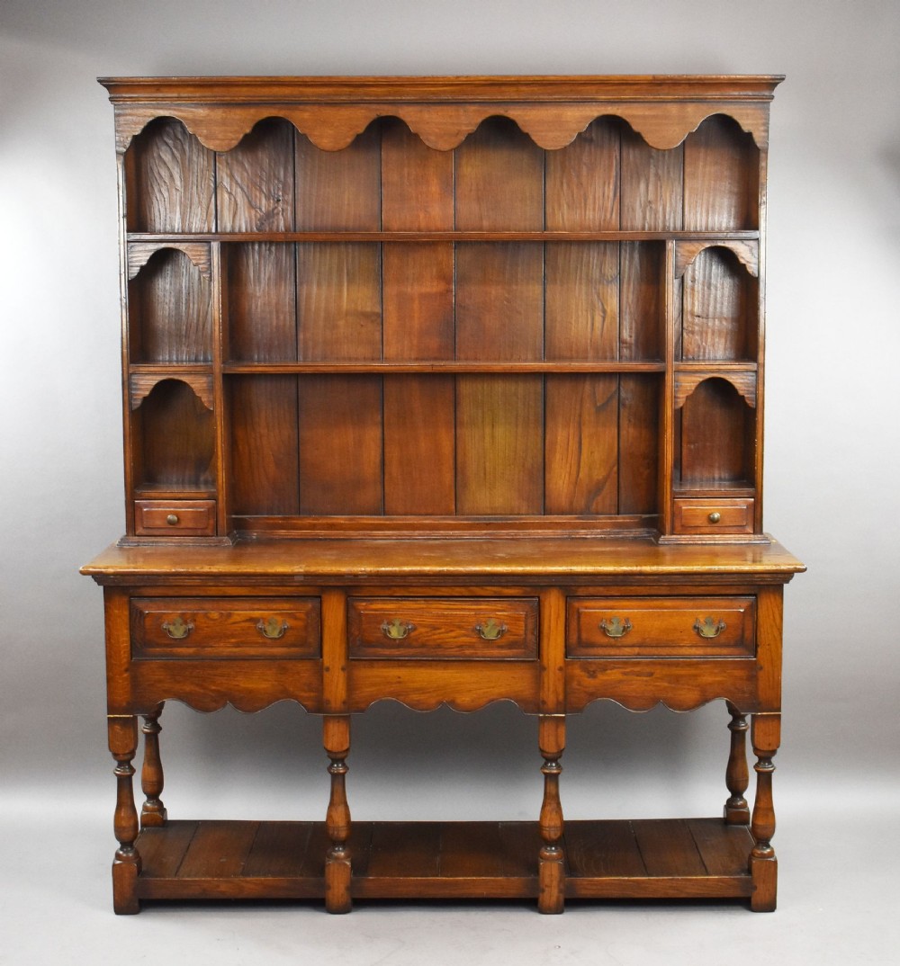 A warm brown oak Welsh dresser featuring an open tiered rack with scalloped aprons above three drawers and a potboard base on turned legs.