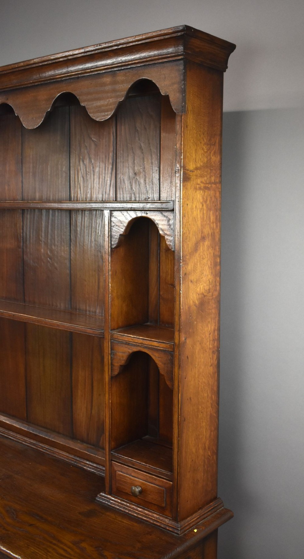 A warm brown oak Welsh dresser featuring an open tiered rack with scalloped aprons above three drawers and a potboard base on turned legs. View VI