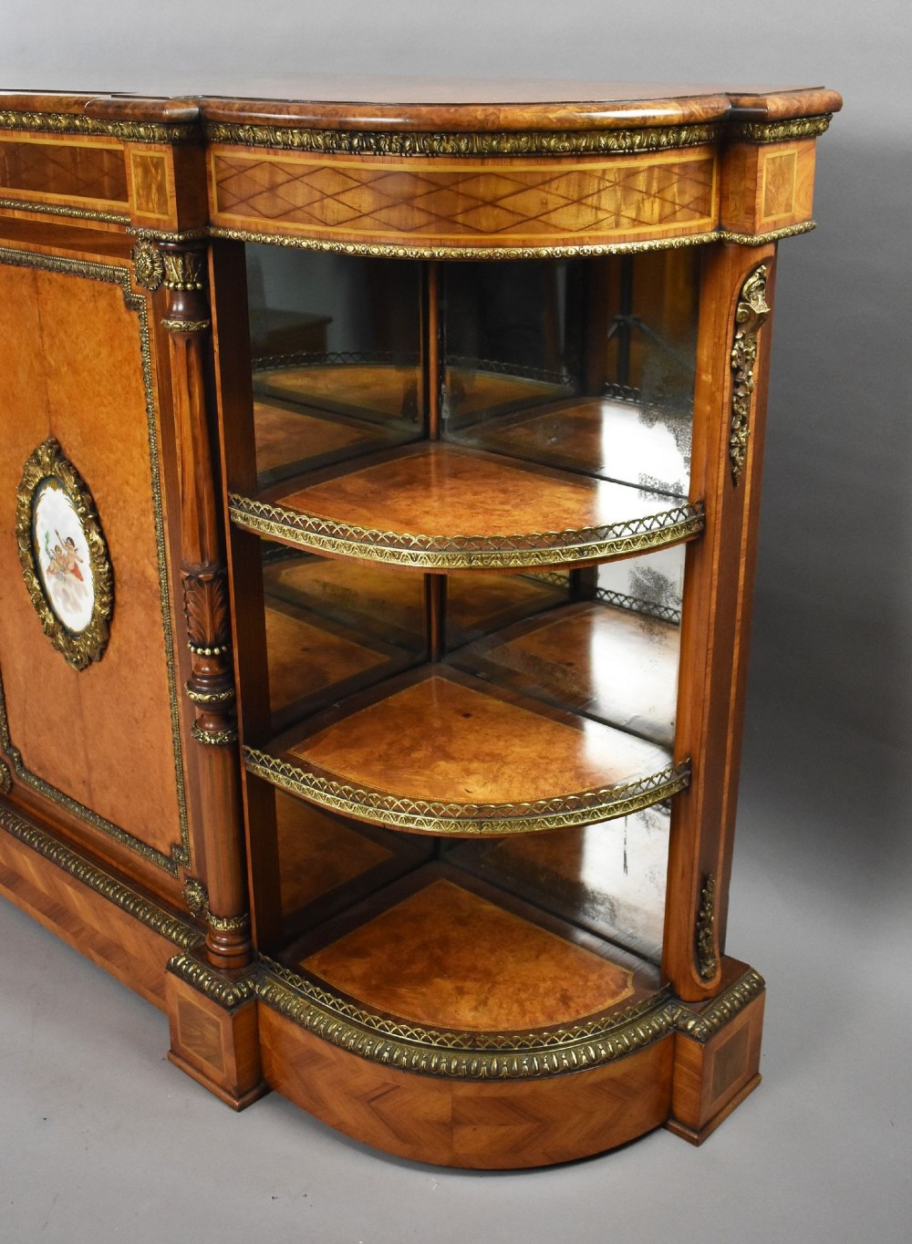 A Victorian burr walnut credenza featuring central cupboard doors with oval porcelain plaques and gilt bronze mounts flanked by curved open display shelves. View X