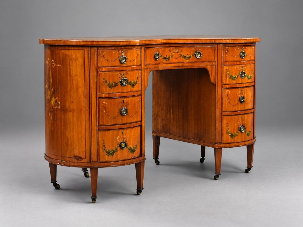 An Edwardian satinwood kidney-shaped writing desk featuring nine hand-painted drawers with brass ring handles and tapered legs on castors.