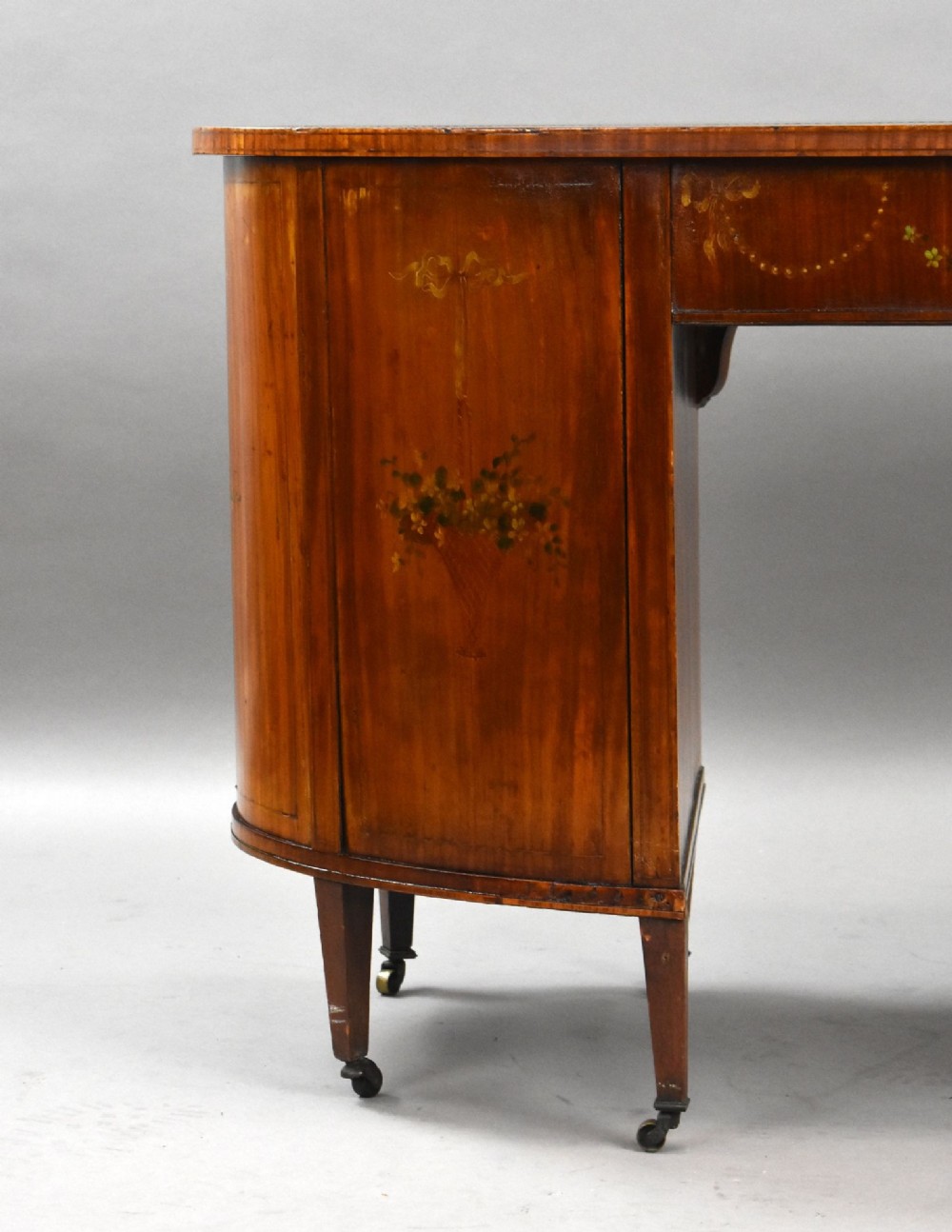 An Edwardian satinwood kidney-shaped writing desk featuring nine hand-painted drawers with brass ring handles and tapered legs on castors. View X