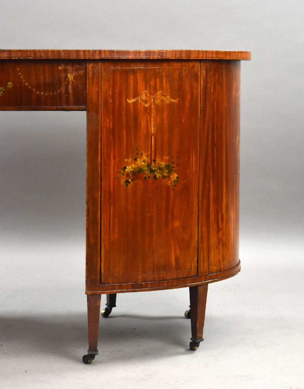An Edwardian satinwood kidney-shaped writing desk featuring nine hand-painted drawers with brass ring handles and tapered legs on castors. View XI