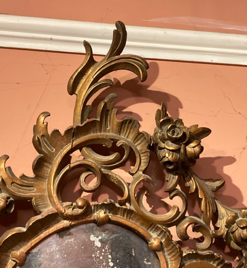 An oval wall mirror featuring an ornate, pierced carved giltwood frame with scrollwork and floral details. View IV
