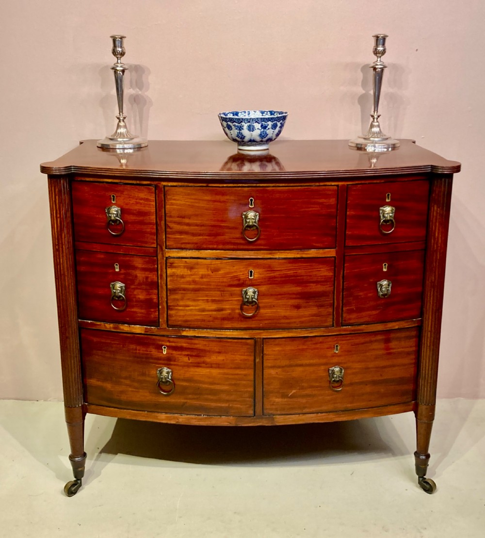 A Regency period warm brown mahogany bow-fronted chest of drawers featuring eight drawers with brass lion-head ring pulls, reeded side pilasters, and turned legs on castors.