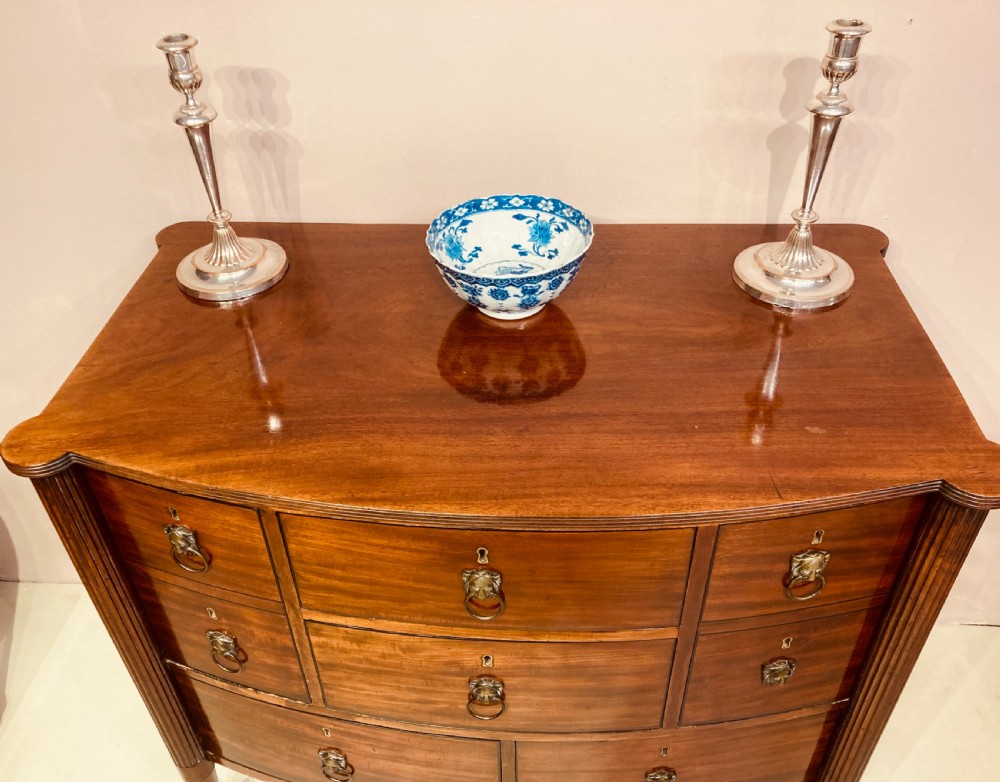 A Regency period warm brown mahogany bow-fronted chest of drawers featuring eight drawers with brass lion-head ring pulls, reeded side pilasters, and turned legs on castors. View II