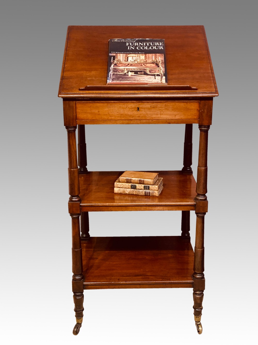 A three-tier Georgian mahogany whatnot with an adjustable slanted reading slope top, a single frieze drawer, turned upright supports, and brass castors.