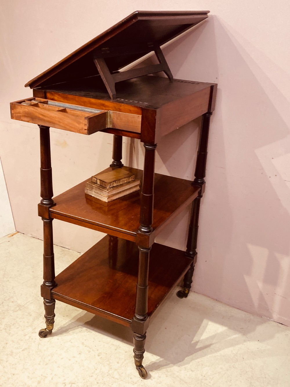A three-tier Georgian mahogany whatnot with an adjustable slanted reading slope top, a single frieze drawer, turned upright supports, and brass castors. View IV