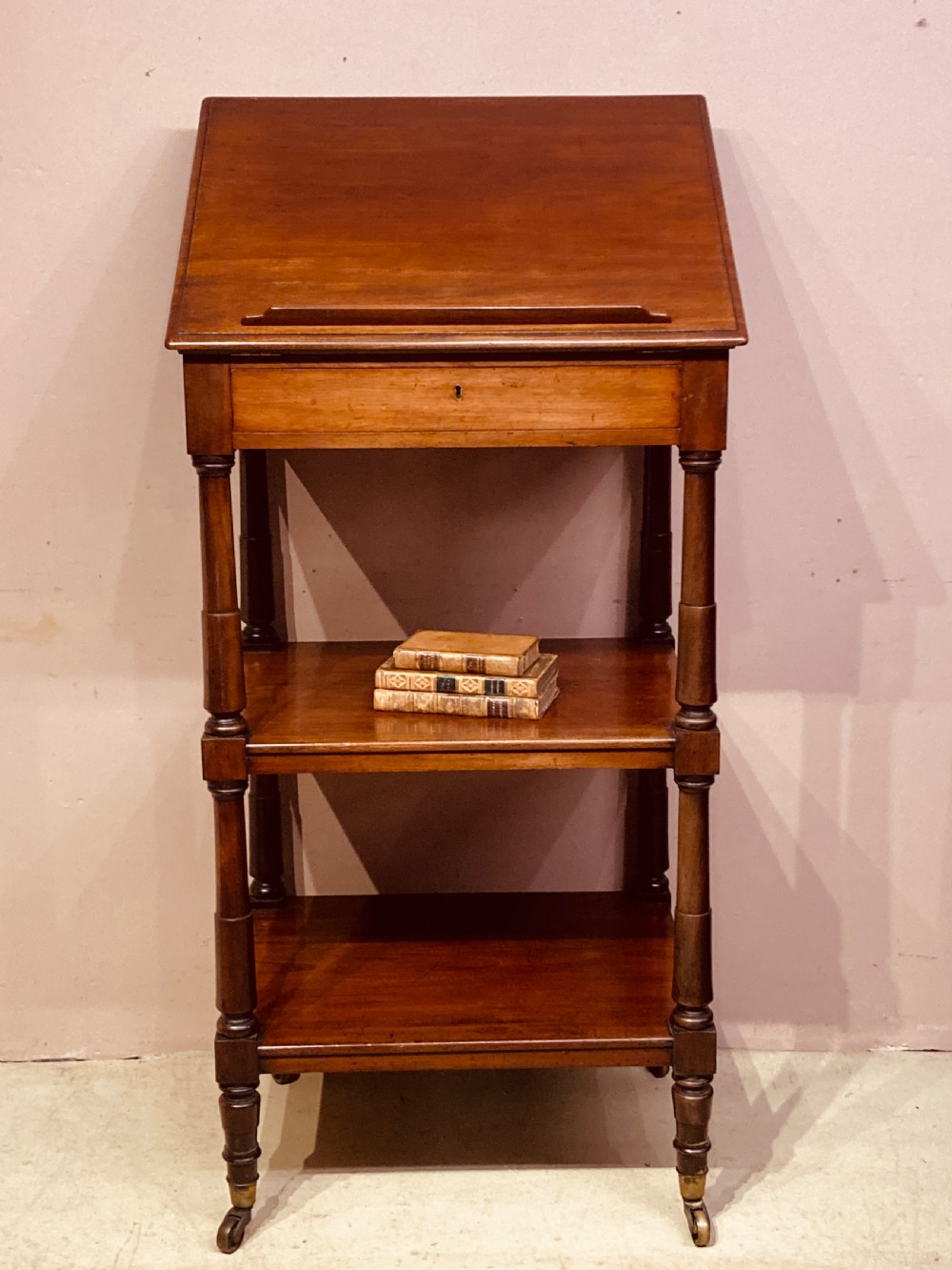 A three-tier Georgian mahogany whatnot with an adjustable slanted reading slope top, a single frieze drawer, turned upright supports, and brass castors. View VI