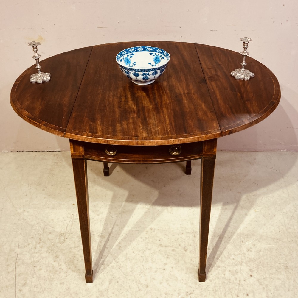 A Georgian mahogany oval Pembroke table featuring extended drop leaves, fine string inlay, a single drawer with brass ring handles, and square tapered legs.