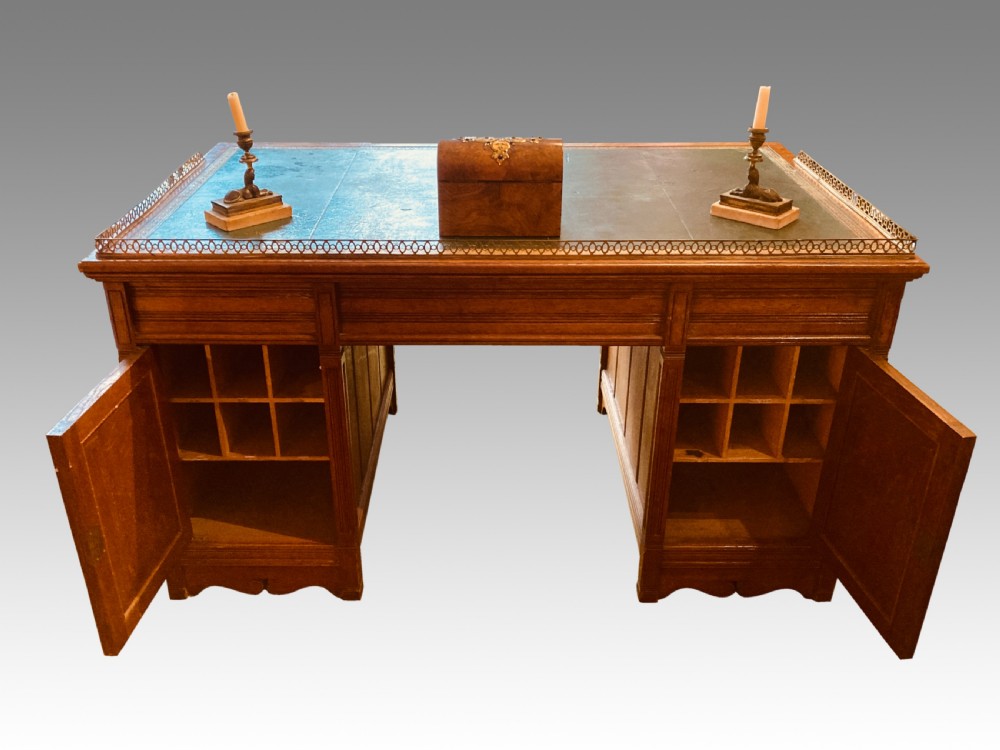 A Victorian oak pedestal desk featuring a green leather writing top, a brass gallery rail, twin banks of drawers with brass ring handles, and a central kneehole. View VI