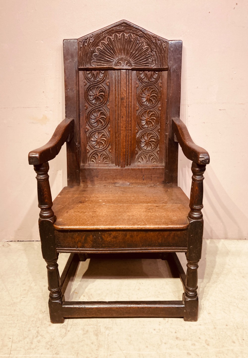 A dark carved oak wainscot armchair featuring an arched cresting rail, carved relief back panel, curved armrests, and turned legs with box stretchers.