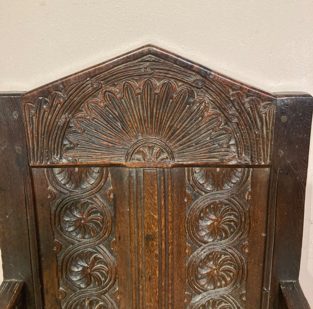 A dark carved oak wainscot armchair featuring an arched cresting rail, carved relief back panel, curved armrests, and turned legs with box stretchers. View II