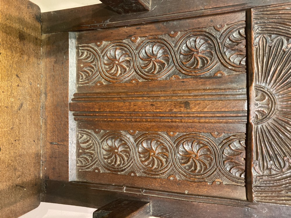 A dark carved oak wainscot armchair featuring an arched cresting rail, carved relief back panel, curved armrests, and turned legs with box stretchers. View III