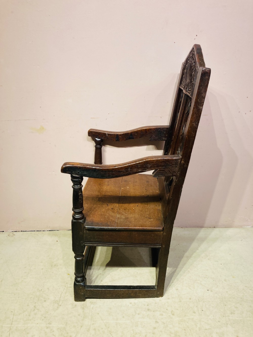 A dark carved oak wainscot armchair featuring an arched cresting rail, carved relief back panel, curved armrests, and turned legs with box stretchers. View VI