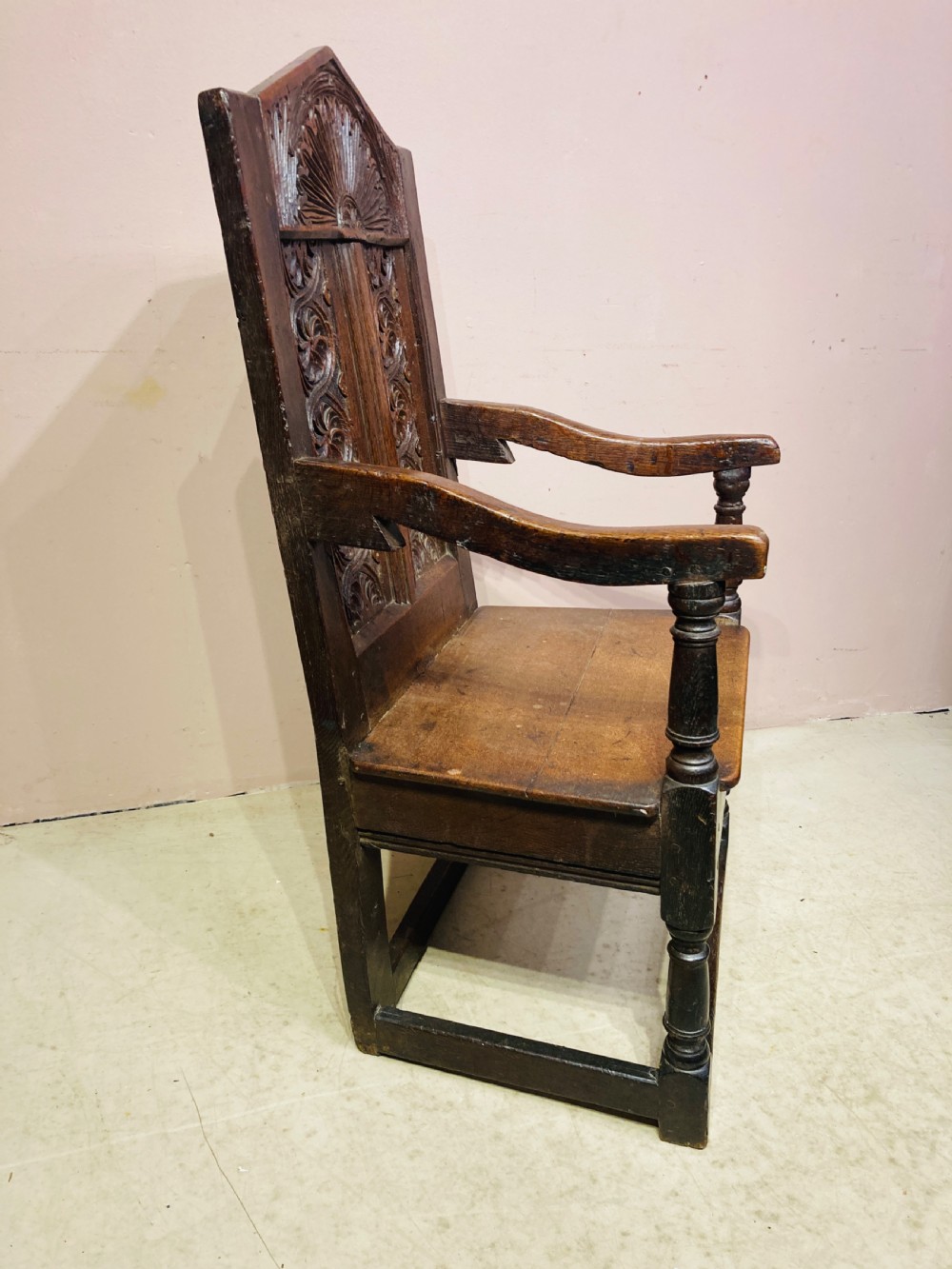 A dark carved oak wainscot armchair featuring an arched cresting rail, carved relief back panel, curved armrests, and turned legs with box stretchers. View IX