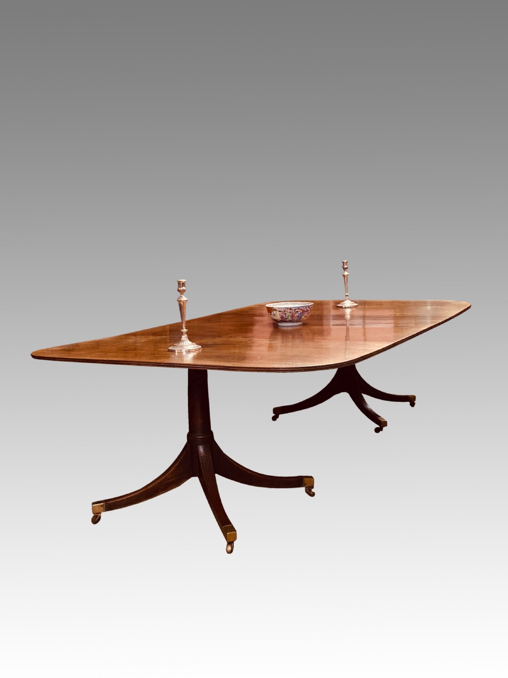 A Georgian mahogany dining table featuring a polished rectangular top with rounded corners supported by two central turned pedestals with splayed reeded legs ending in brass castors.