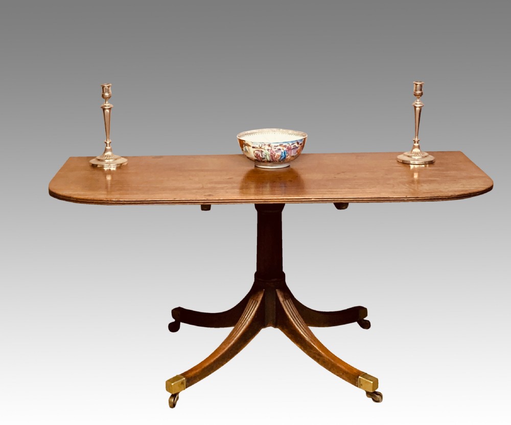 A Georgian mahogany dining table featuring a polished rectangular top with rounded corners supported by two central turned pedestals with splayed reeded legs ending in brass castors. View VI