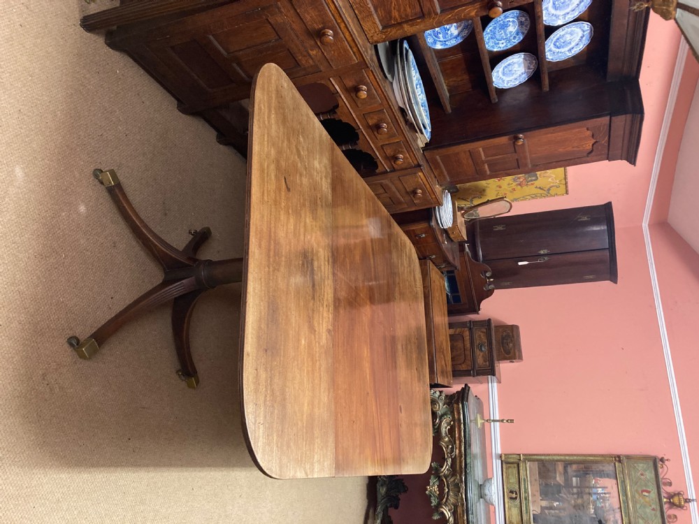 A Georgian mahogany dining table featuring a polished rectangular top with rounded corners supported by two central turned pedestals with splayed reeded legs ending in brass castors. View IX