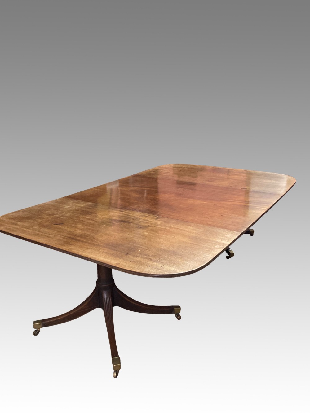 A Georgian mahogany dining table featuring a polished rectangular top with rounded corners supported by two central turned pedestals with splayed reeded legs ending in brass castors. View X