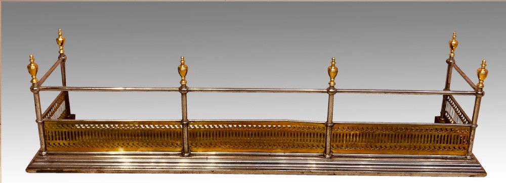 An antique polished steel hearth fender featuring a pierced brass lower gallery band, vertical support posts, and five turned brass urn finials along the top rail.