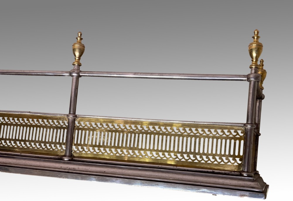 An antique polished steel hearth fender featuring a pierced brass lower gallery band, vertical support posts, and five turned brass urn finials along the top rail. View II
