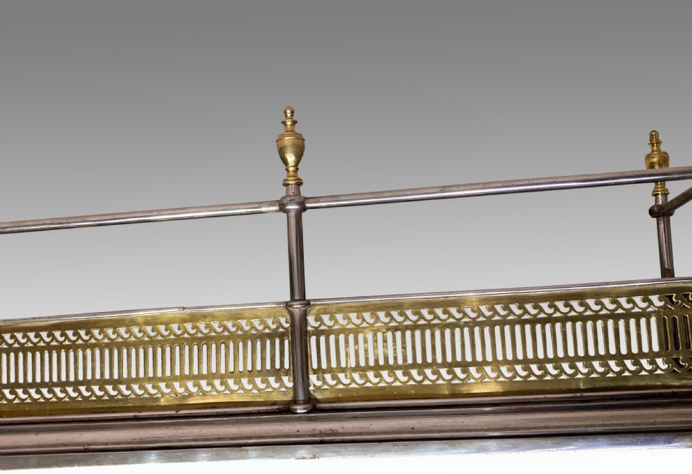An antique polished steel hearth fender featuring a pierced brass lower gallery band, vertical support posts, and five turned brass urn finials along the top rail. View V