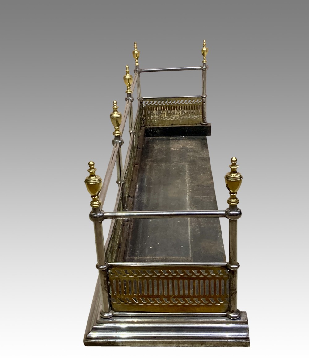 An antique polished steel hearth fender featuring a pierced brass lower gallery band, vertical support posts, and five turned brass urn finials along the top rail. View VI