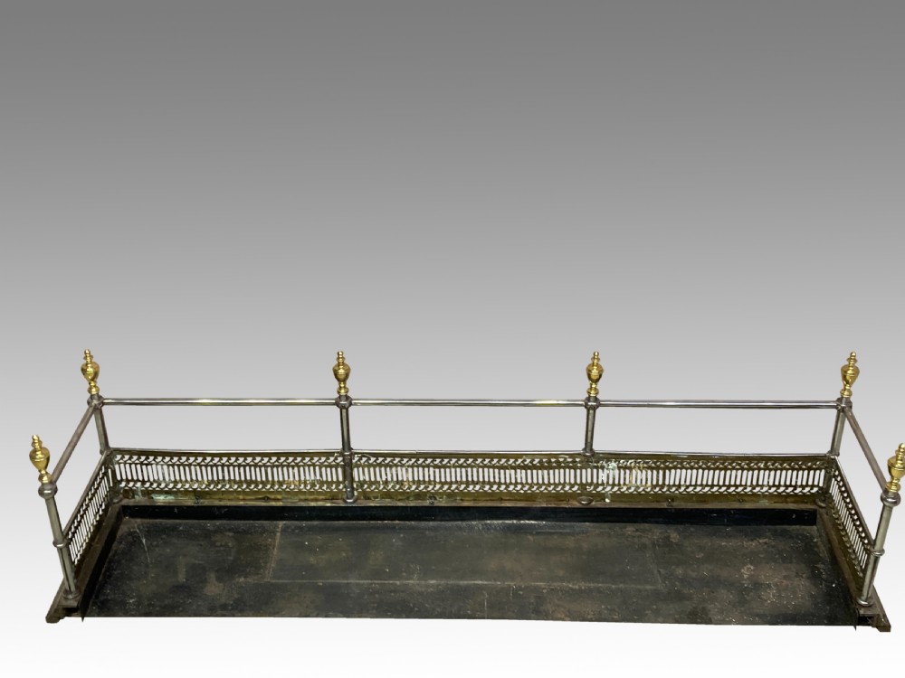 An antique polished steel hearth fender featuring a pierced brass lower gallery band, vertical support posts, and five turned brass urn finials along the top rail. View VII