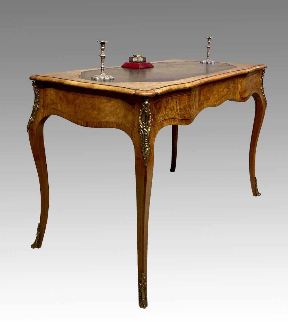 A Victorian burr walnut writing table featuring cabriole legs, ornate gilt-bronze mounts, a serpentine apron, and a dark leather inset writing surface.