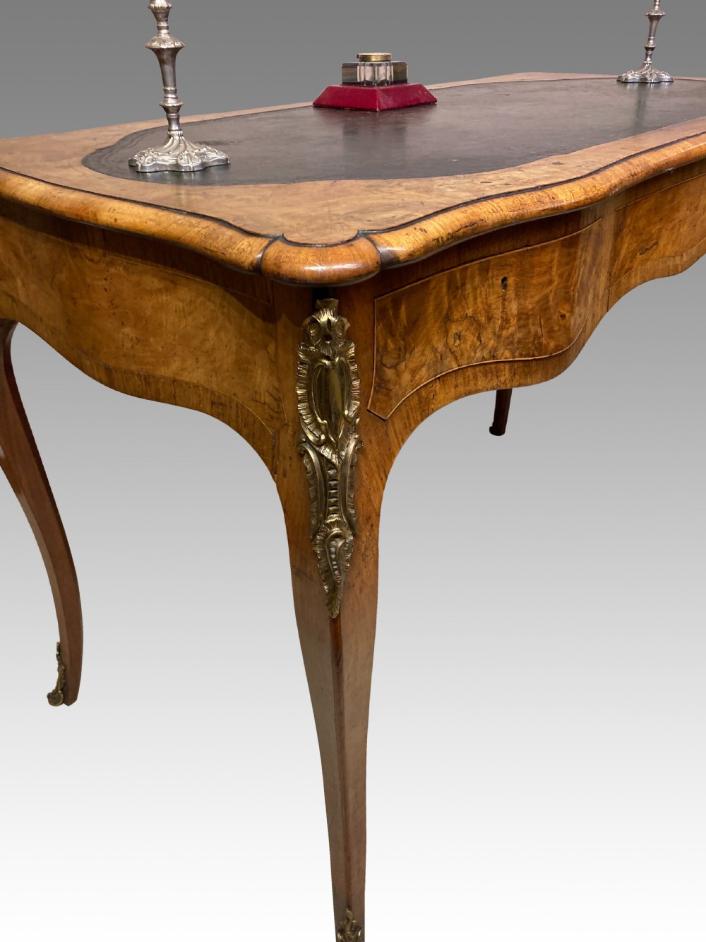 A Victorian burr walnut writing table featuring cabriole legs, ornate gilt-bronze mounts, a serpentine apron, and a dark leather inset writing surface. View IV