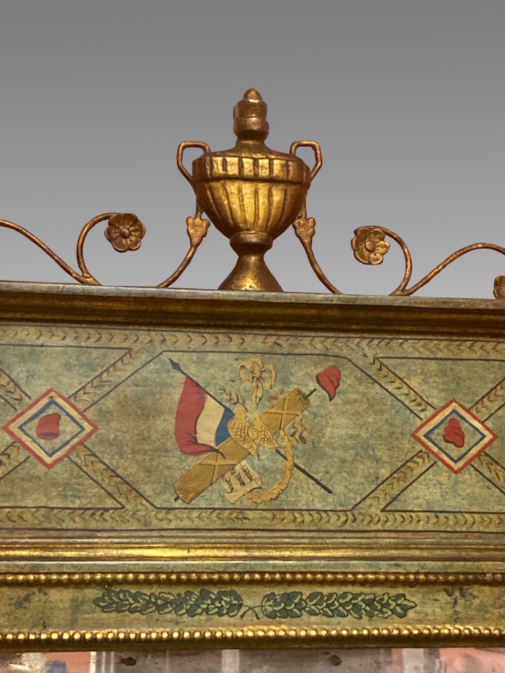 A tall Neoclassical French pier mirror featuring a green painted frame, gilded beaded trim, an urn motif cresting, and two hand-painted decorative panels. View II