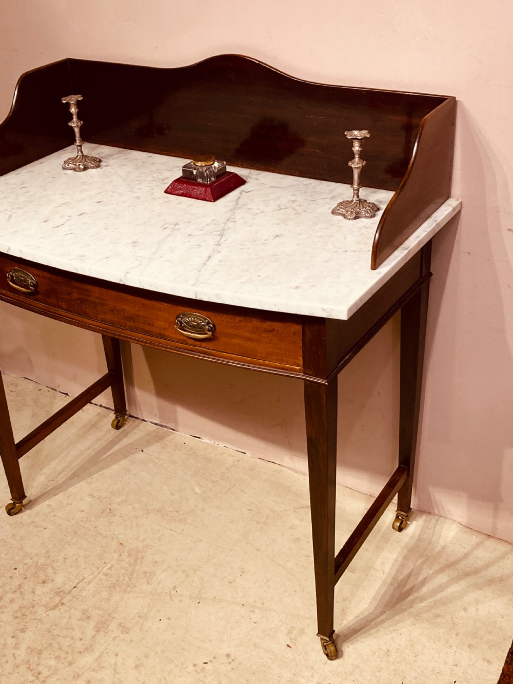 A 19th-century mahogany bow-fronted side table featuring a white marble top, a wooden gallery back, a single drawer with brass handles, and slender tapered legs on castors. View IV