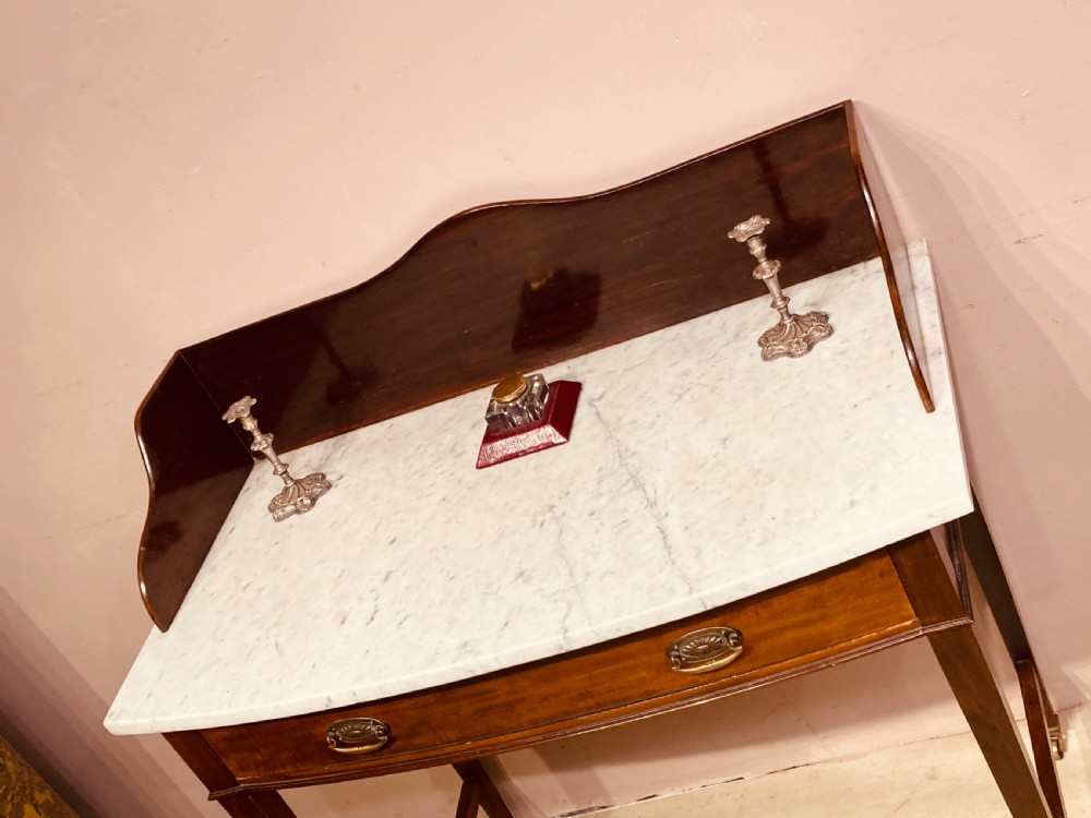 A 19th-century mahogany bow-fronted side table featuring a white marble top, a wooden gallery back, a single drawer with brass handles, and slender tapered legs on castors. View V