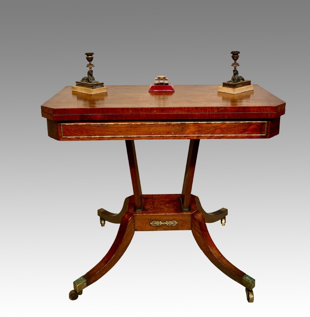A Regency rosewood fold-over card table featuring brass inlay, a central pedestal support, and four curved sabre legs on brass castors.