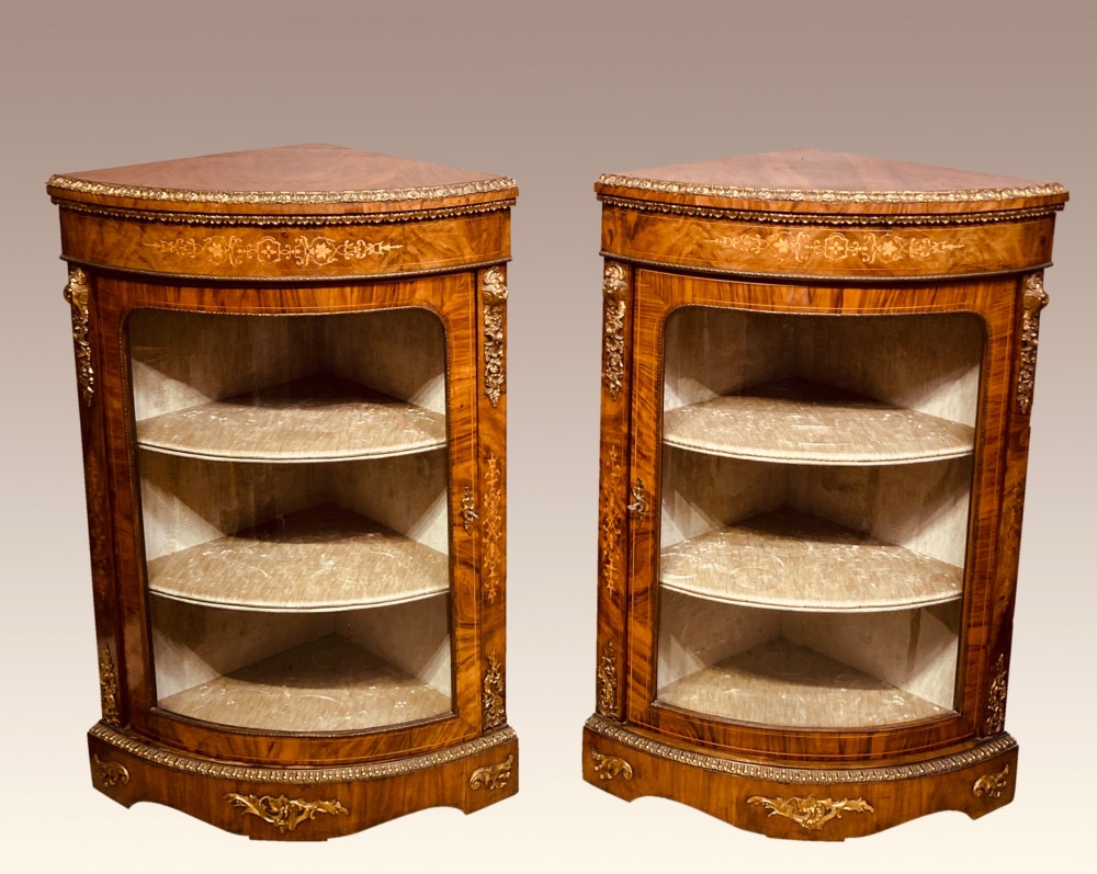 A pair of Victorian inlaid walnut corner cabinets featuring curved glass display doors, decorative floral marquetry, and gilt bronze mounts.