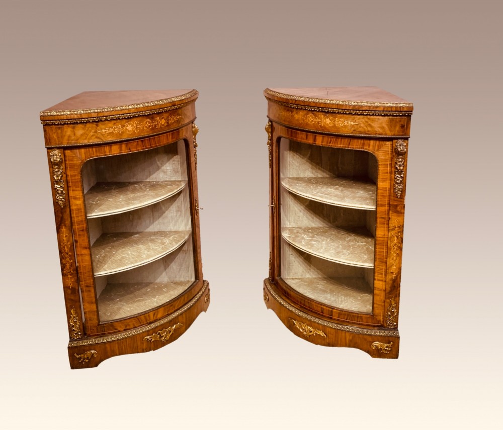 A pair of Victorian inlaid walnut corner cabinets featuring curved glass display doors, decorative floral marquetry, and gilt bronze mounts. View VI