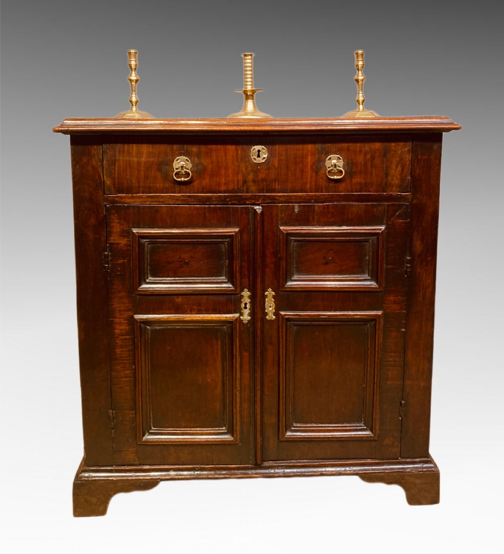 An antique dark brown walnut cupboard with a single top drawer, two fielded panel doors, brass drop handles, and bracket feet.