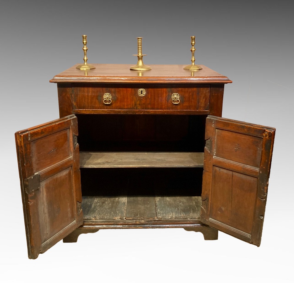 An antique dark brown walnut cupboard with a single top drawer, two fielded panel doors, brass drop handles, and bracket feet. View III