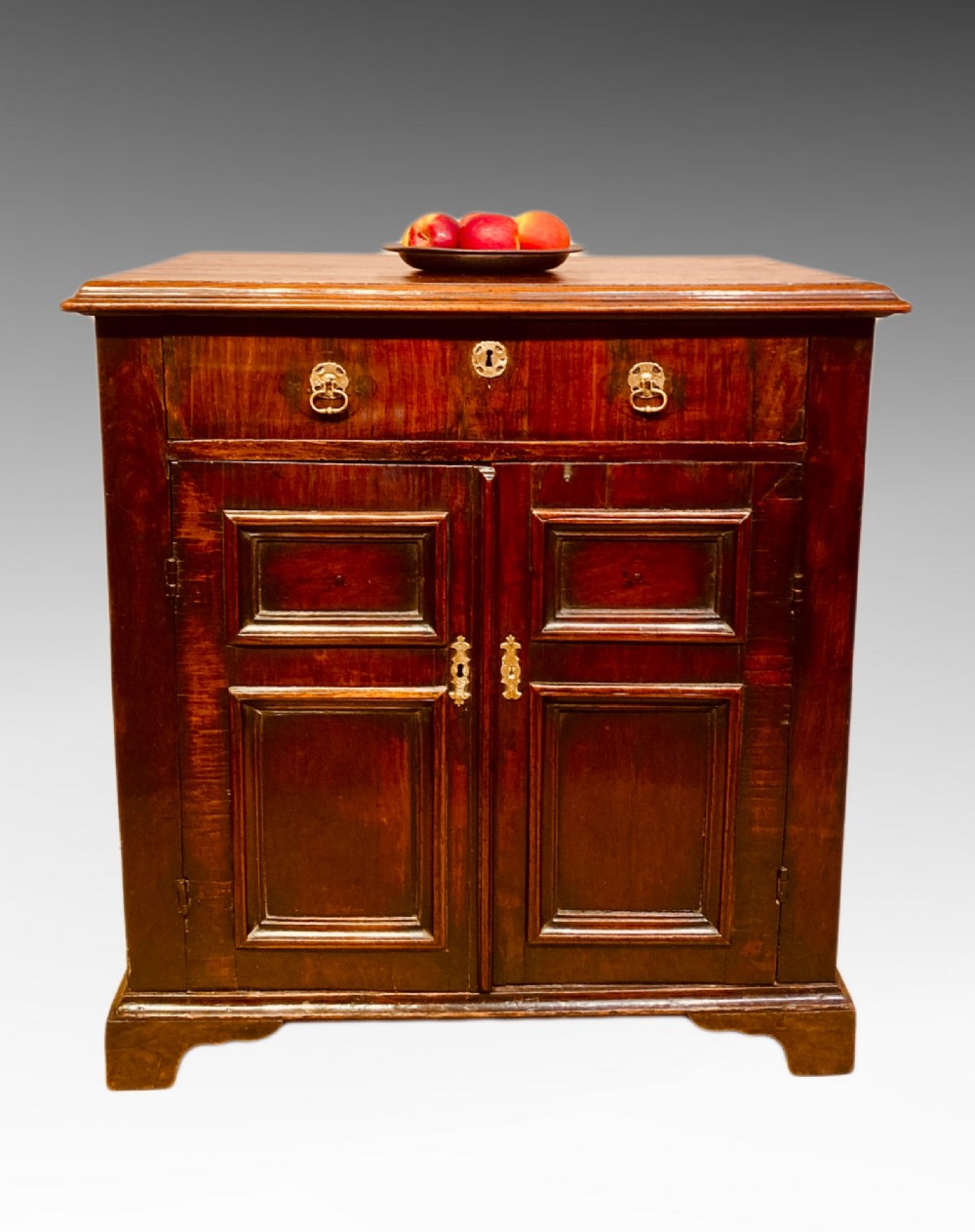 An antique dark brown walnut cupboard with a single top drawer, two fielded panel doors, brass drop handles, and bracket feet. View V