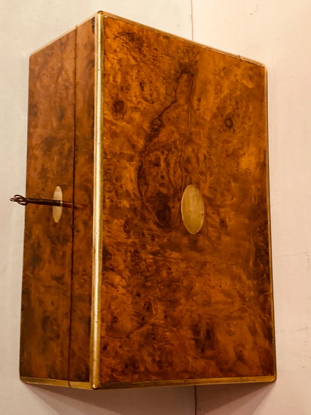 A rectangular Late Georgian burr walnut writing slope box featuring brass edging, an oval brass lid plaque, and a keyhole with key on the front. View II