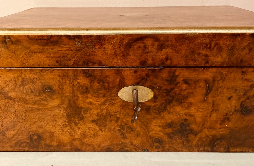A rectangular Late Georgian burr walnut writing slope box featuring brass edging, an oval brass lid plaque, and a keyhole with key on the front. View X