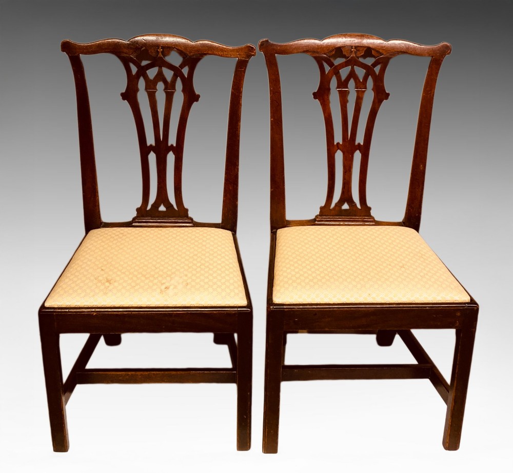 A pair of antique Georgian mahogany side chairs featuring pierced Gothic splat backs, cream upholstered seats, and straight legs connected by stretchers.